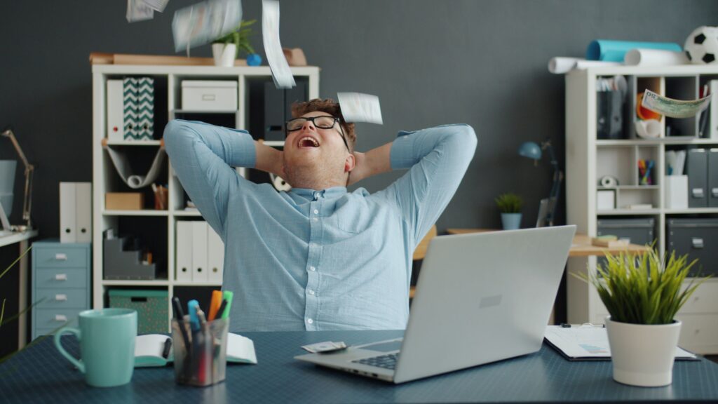 pexels photo 36764805 36764805 A happy businessman relaxes at his desk as banknotes fall around him, symbolizing success.